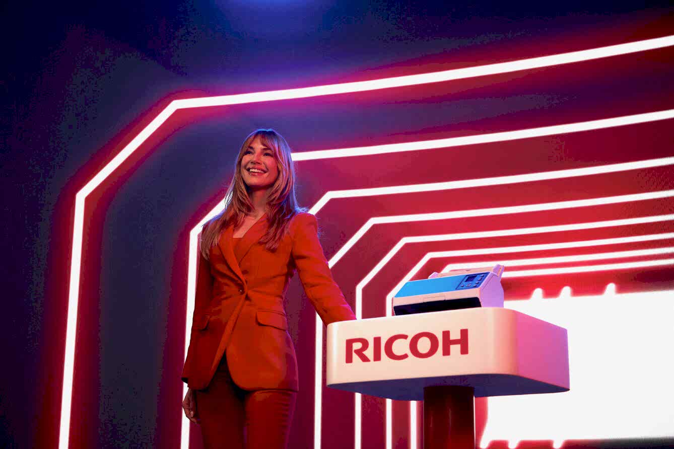 Smiling presenter in a red suit standing at a RICOH branded podium with neon lighting in the background