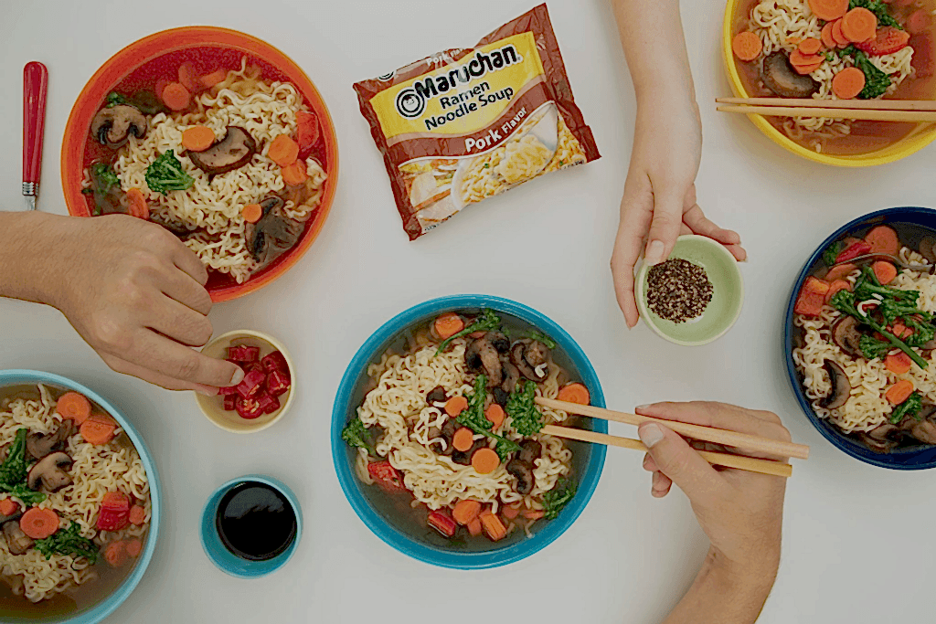 Hands preparing and serving instant ramen noodles with vegetables and pork flavor packet visible