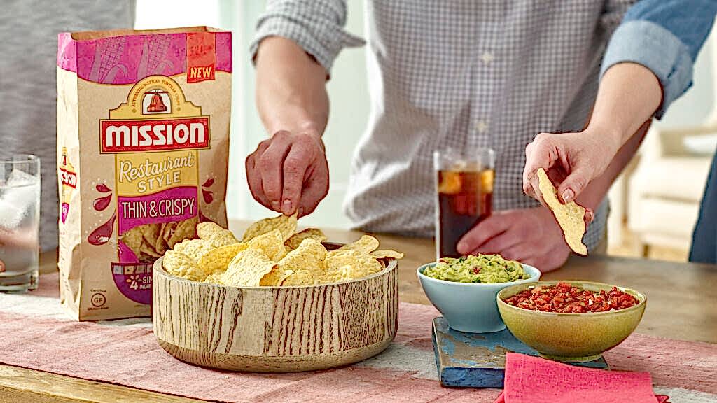 Hands serving Mission tortilla chips from a bowl with guacamole and salsa on the side