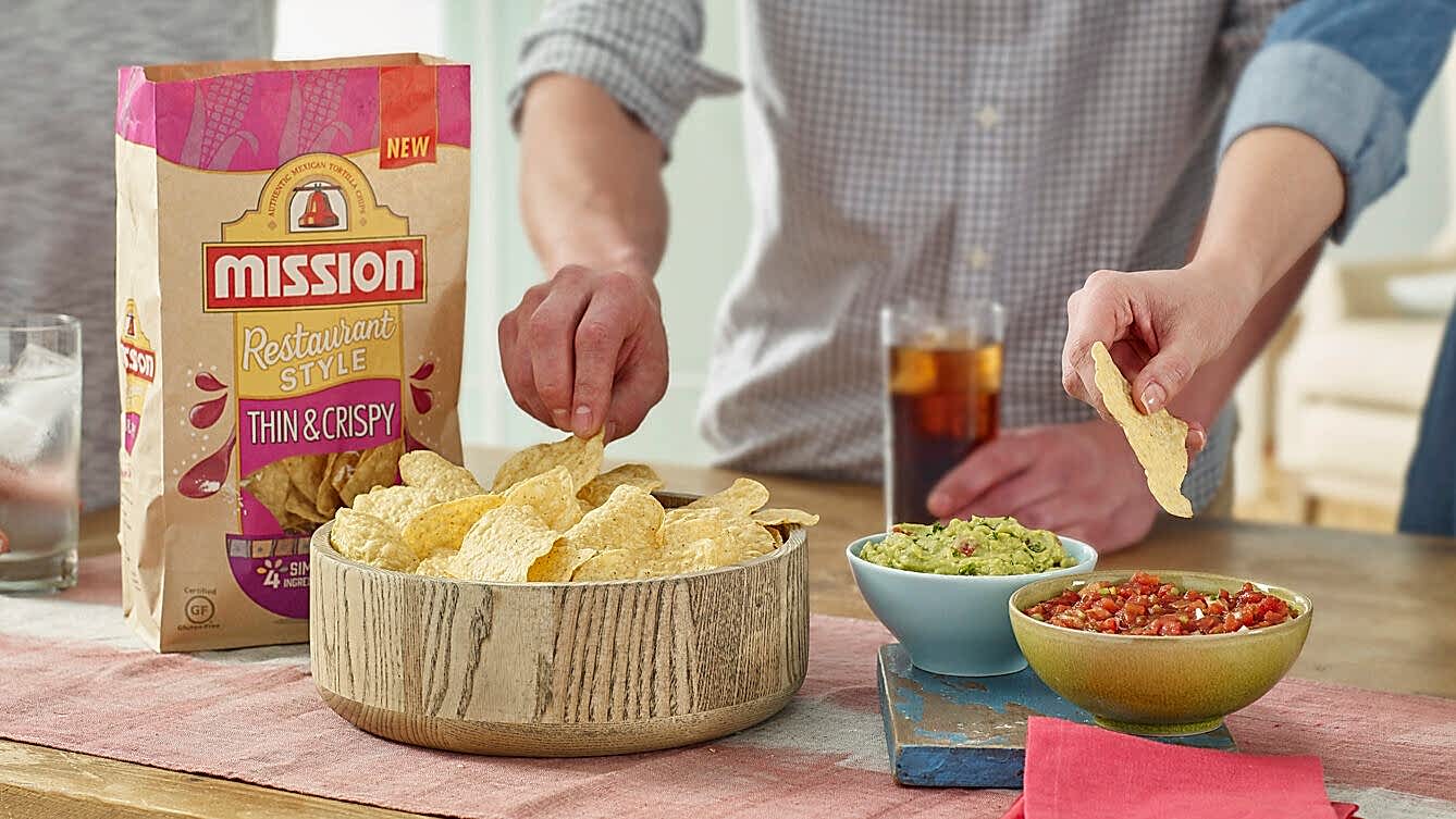 Hands serving Mission tortilla chips from a bowl with guacamole and salsa on the side