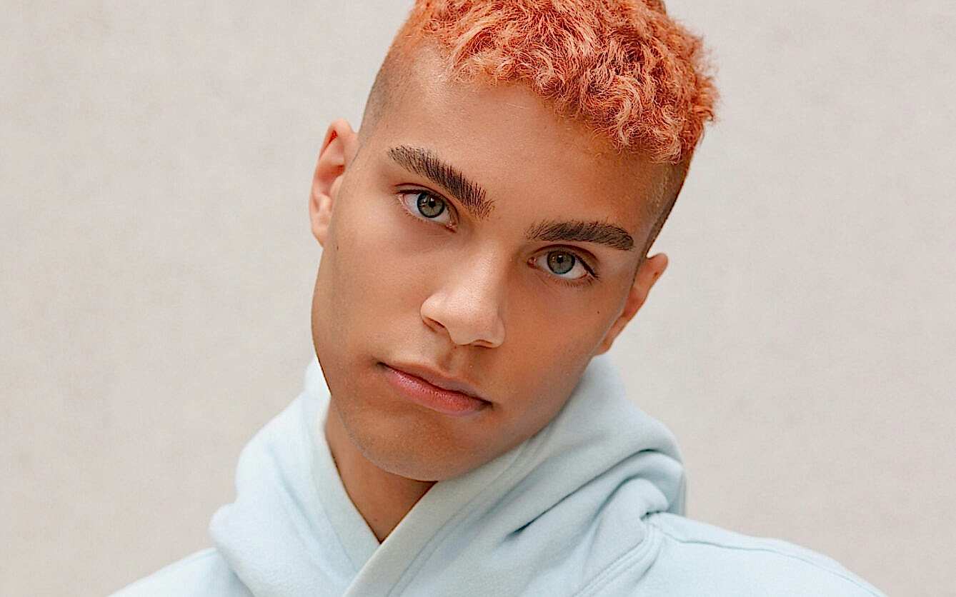 This is a close-up of a young man with peach-colored buzz-cut hair and clear hazel eyes. He has well-groomed eyebrows and a neutral expression and is wearing a light blue hoodie. The background is simple and muted, focusing attention on his face.