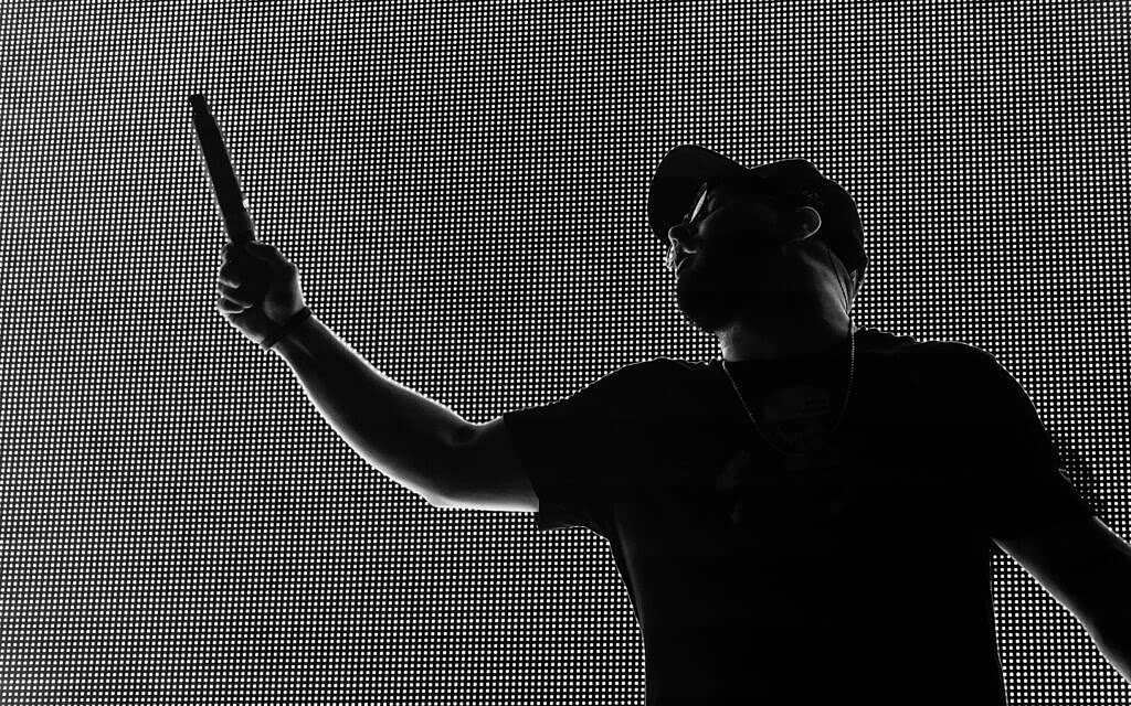 Silhouette of a man against a bright LED screen, raising his right hand and pointing upwards with a microphone in hand. He wears a cap, a T-shirt, and a necklace. The background LED screen creates a high-contrast pixelated effect around his outline.