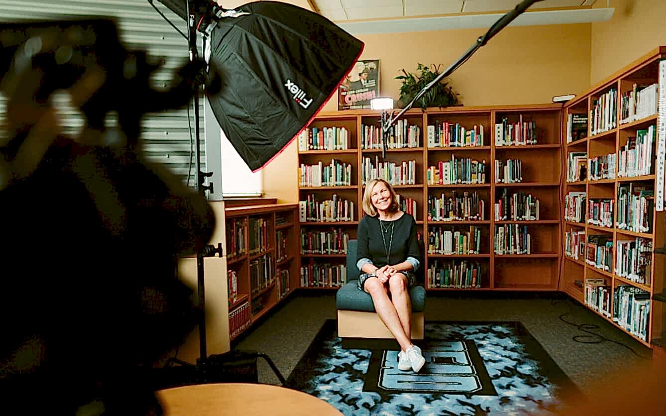 A woman sits smiling on a chair in a library for a video interview, with a camera and lighting equipment directed toward her. She is casually dressed in a dark blue dress and sneakers, with crossed legs and folded hands. The background is filled with bookshelves stocked with colorful books, and the ambiance suggests a candid, professional setting.