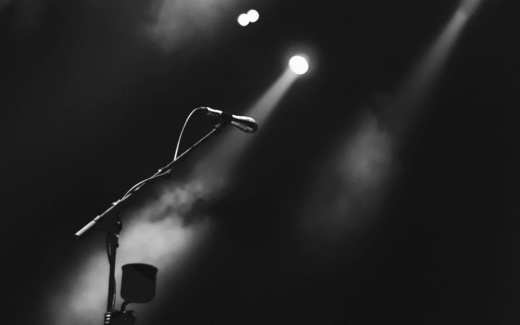 empty stage with a single microphone stand illuminated by a beam of light against a dark background.