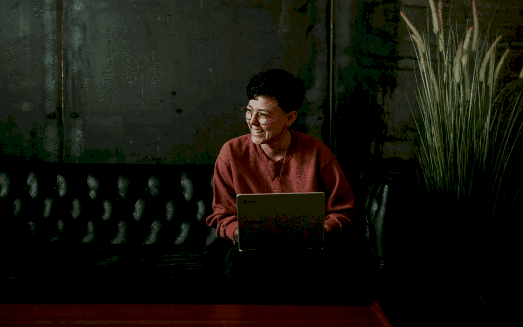 Person in a red sweater and glasses smiling and looking off to the side while sitting on a black leather couch with a laptop on their lap, in a room with a concrete wall and a large potted grass plant