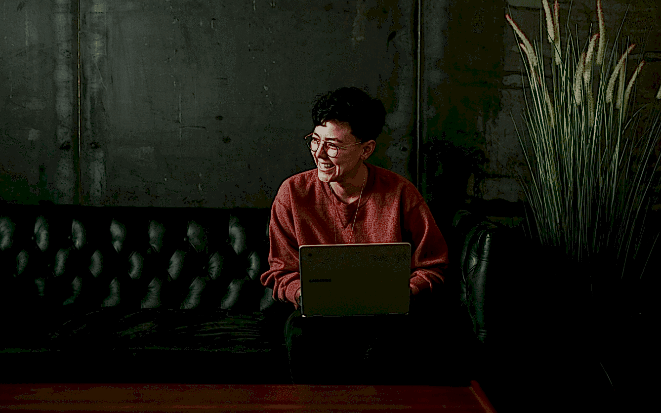 Person in a red sweater and glasses smiling and looking off to the side while sitting on a black leather couch with a laptop on their lap, in a room with a concrete wall and a large potted grass plant