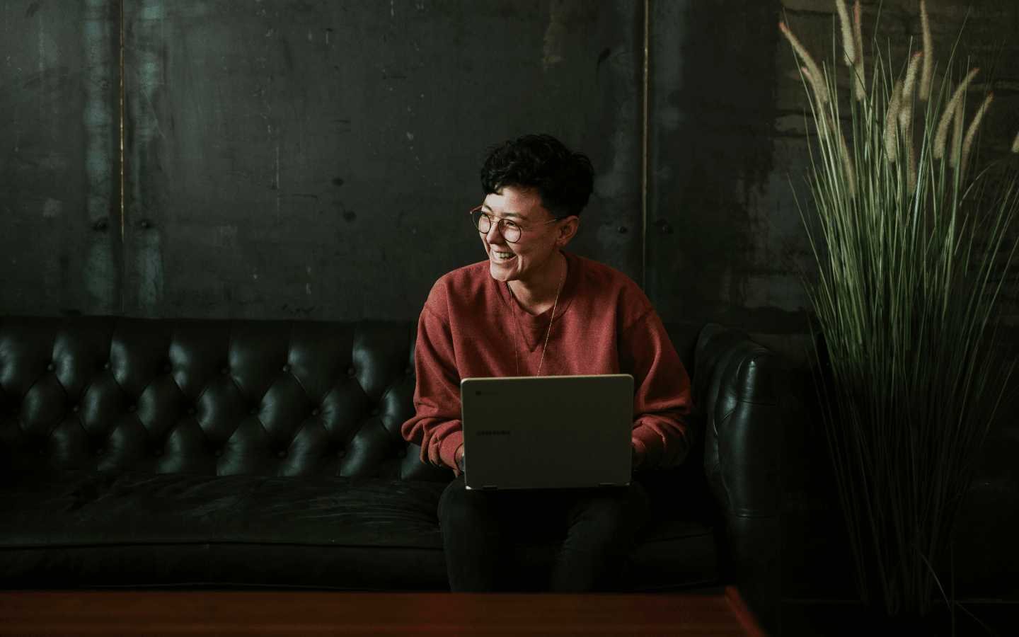 Person in a red sweater and glasses smiling and looking off to the side while sitting on a black leather couch with a laptop on their lap, in a room with a concrete wall and a large potted grass plant