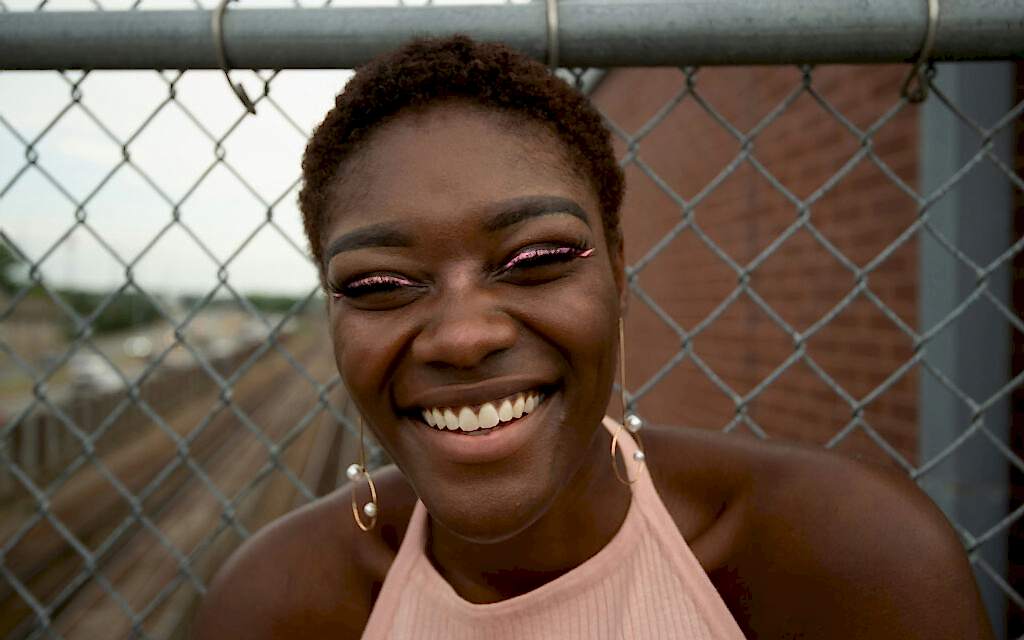 A young woman with a radiant smile, sporting short curly hair, hoop earrings, and glittery eyeliner, leans against a chain-link fence with an urban backdrop.