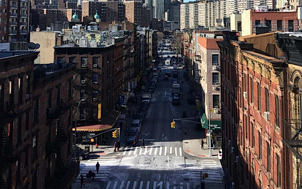 "Elevated view of a bustling city street intersection with pedestrian crosswalks, traffic lights, and rows of classic brick buildings on either side, under a clear blue sky.