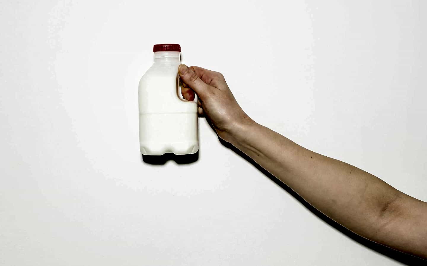 A person's arm is extended towards the camera. The person is holding a small plastic milk bottle with a red cap against a plain white background.