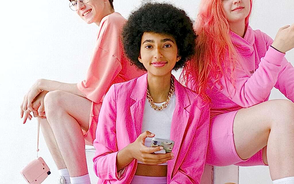 Three young women in varying shades of pink attire posing casually. The central figure, holding a smartphone, wears a pink blazer and has a curly afro, while the other two, one with glasses and the other with long pink hair, appear slightly in the background, all against a plain white backdrop.