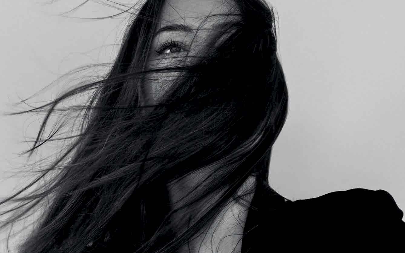 A black-and-white close-up portrait of a woman with her face partially obscured by her flowing dark hair creates a mysterious and artistic effect.