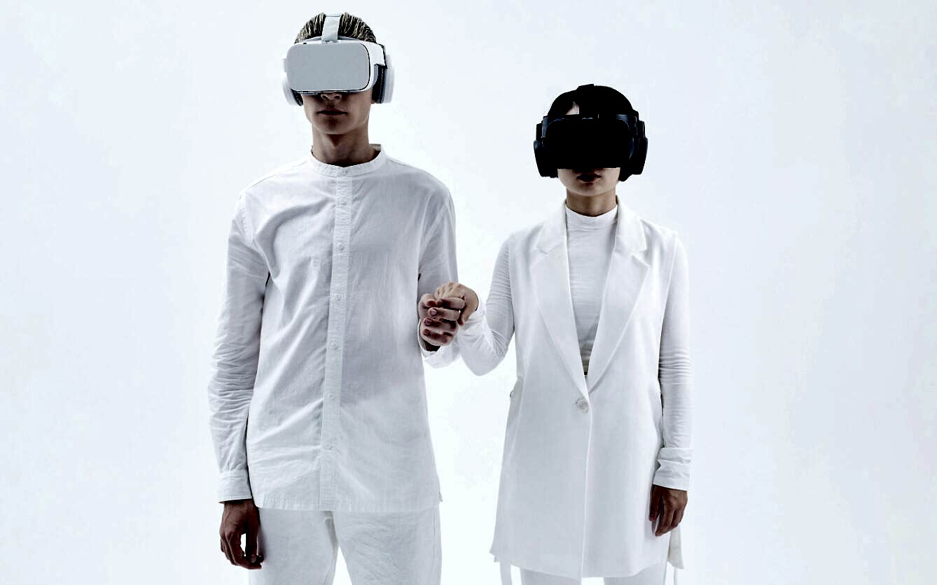 Two people wearing VR headsets and holding hands, dressed in white outfits, standing against a plain white background.