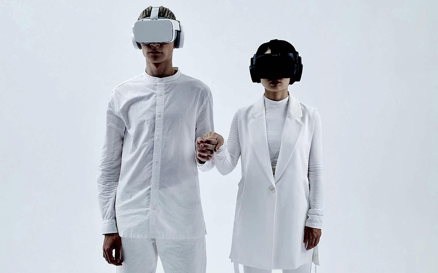 Two people wearing VR headsets and holding hands, dressed in white outfits, standing against a plain white background.