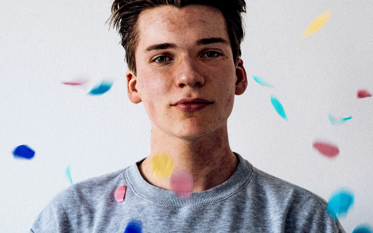 Young person with short hair wearing a gray shirt, with colorful confetti floating around, against a plain white background.