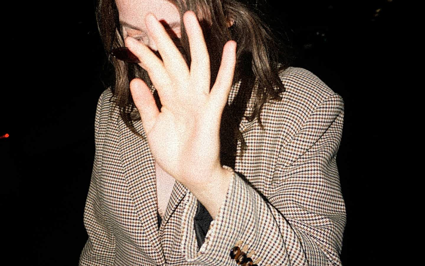 A person with long hair wearing a plaid blazer, raising their hand to cover their face against a dark background.