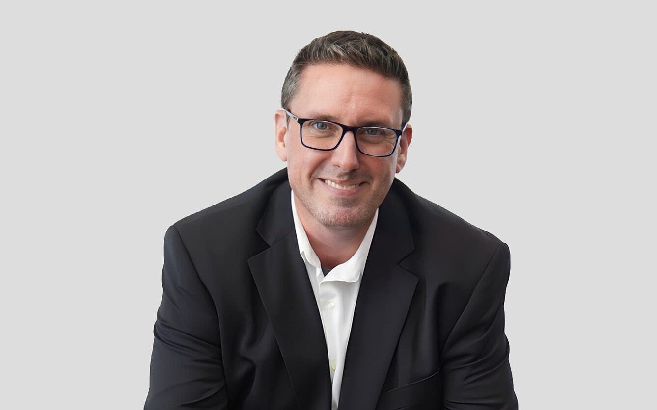 A smiling man in a black suit and white shirt wearing glasses is sitting against a plain grey background.