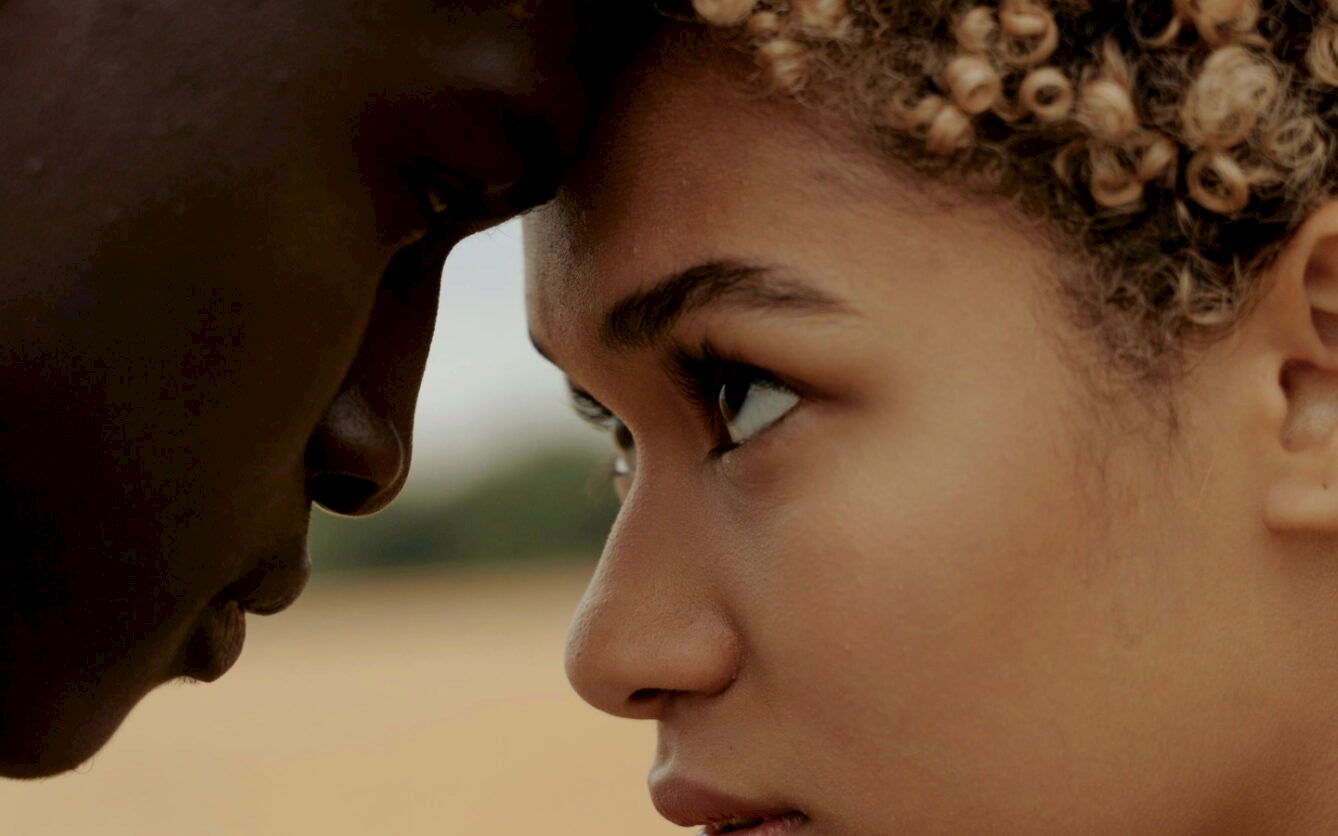 This is a close-up image of a young woman and a man touching foreheads, showcasing an intimate moment. The focus is on the woman's profile, capturing her detailed facial features and expressive eyes set against a blurred natural background.