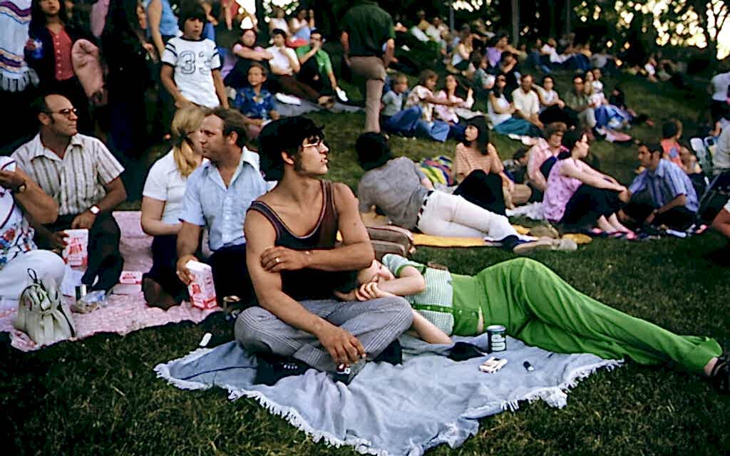 This is a vintage scene of diverse people relaxing on a grassy hill at a park during a public event. It features a man in striped pants and a tank top reclining with a camera, with others lounging and conversing around blankets spread out with snacks and drinks.