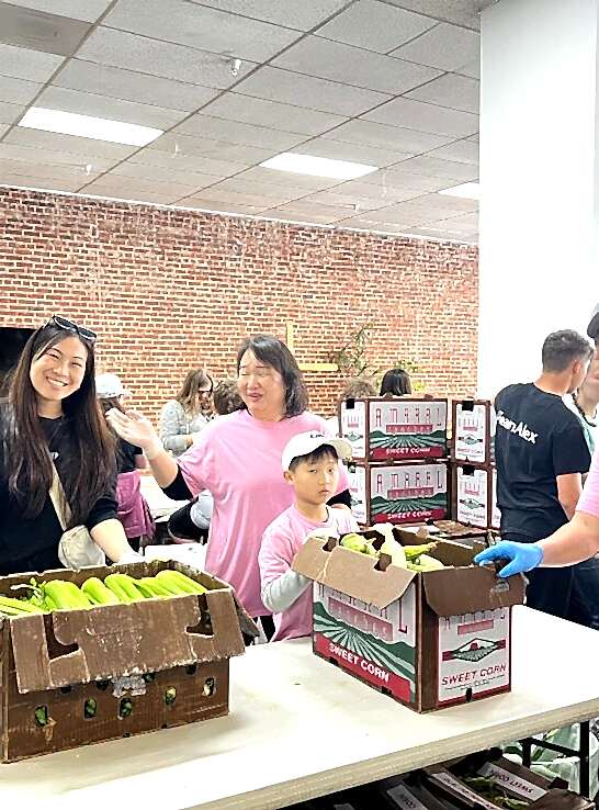 Volunteers, including a woman and child, smiling while packing corn in cardboard boxes at a community service event