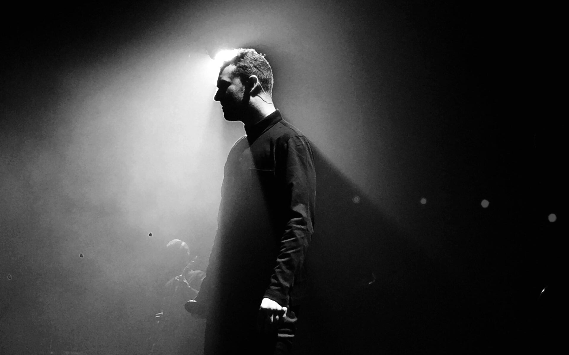 Black and white image of a person standing in a spotlight, silhouetted against a dark background. The dramatic lighting creates a strong contrast, emphasizing the figure’s profile and the atmospheric, moody setting.