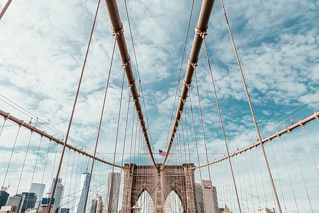 View of the Brooklyn Bridge cables and towers with an American flag against a cloudy blue sky and the New York City skyline in the background